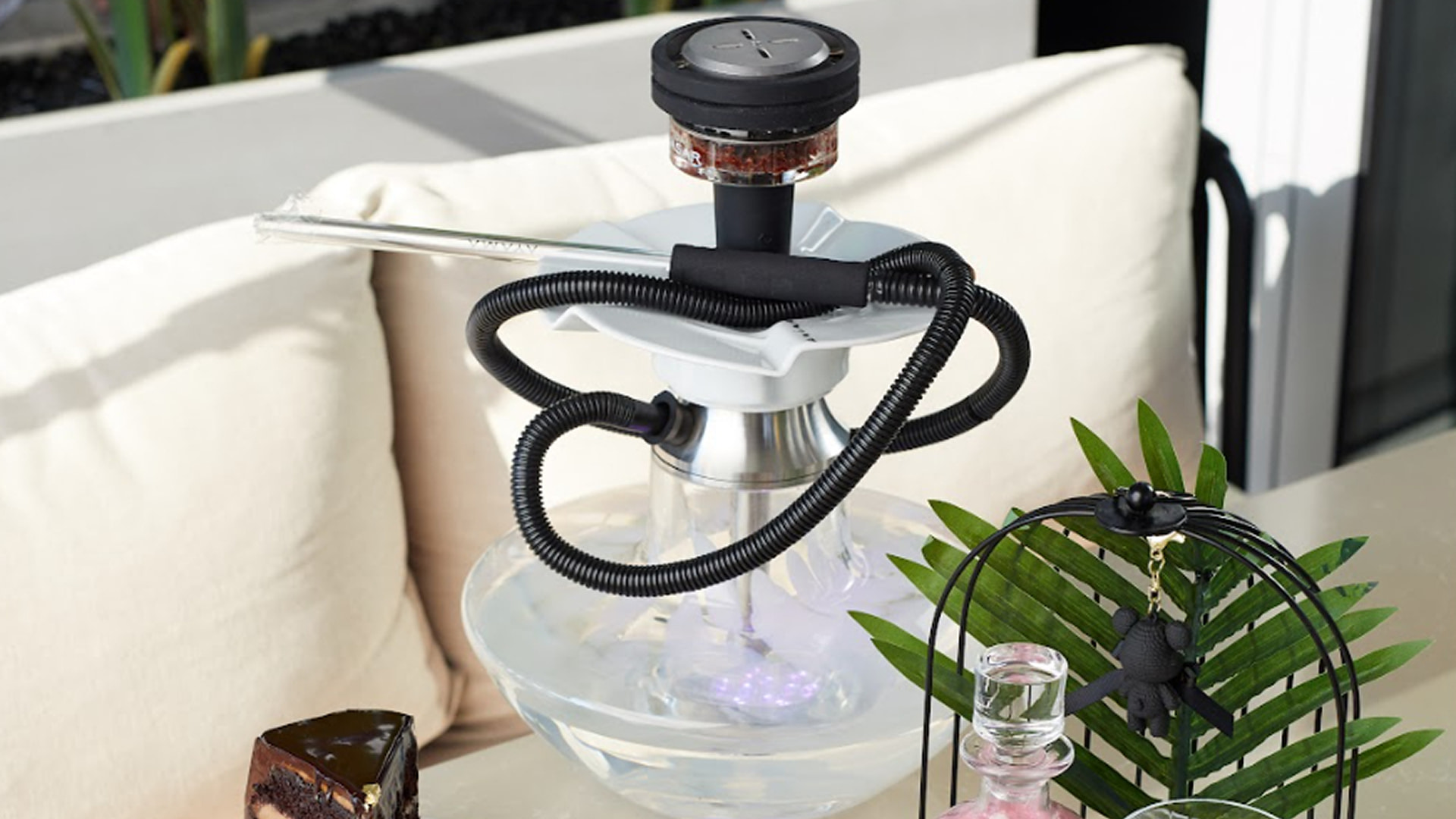 Modern Russian shisha setup with glass base, metal stem, and hose placed on a stylish outdoor lounge table with cushions.