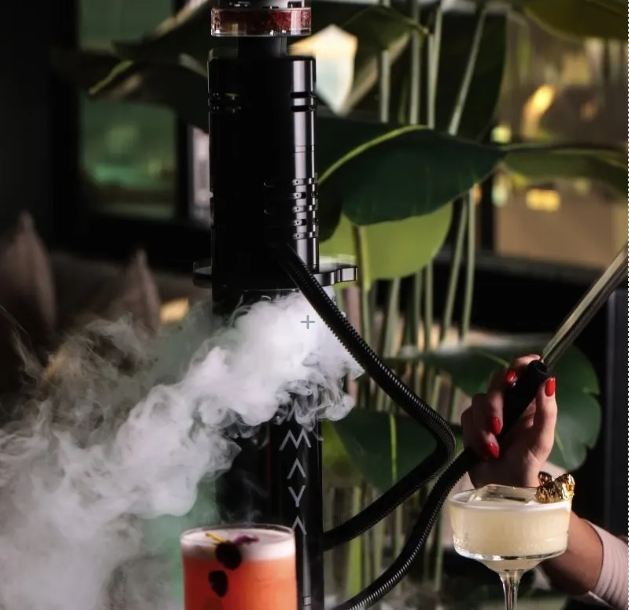 A black shisha pipe releases a cloud of smoke beside two cocktails, with a hand holding the hose in a dim, modern lounge setting