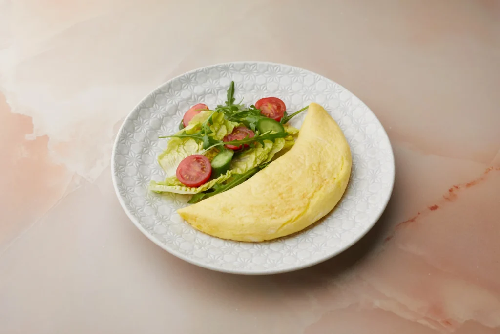 Fluffy folded omelette served with a fresh side salad of lettuce, cherry tomatoes, cucumber, and arugula on a patterned white plate at AMAYA, Dubai Mall.