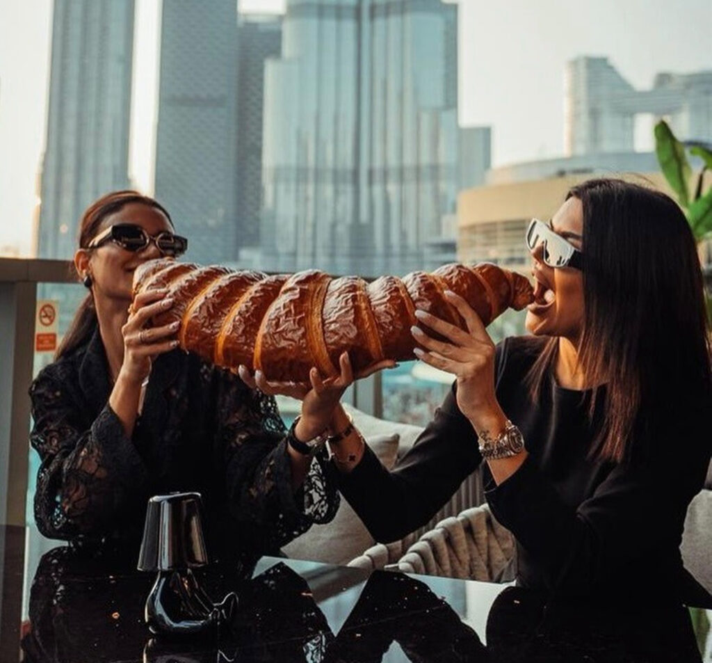 Two stylish individuals in chic attire share an indulgent oversized croissant at a glossy café table, enjoying a high‑end breakfast with panoramic city views.