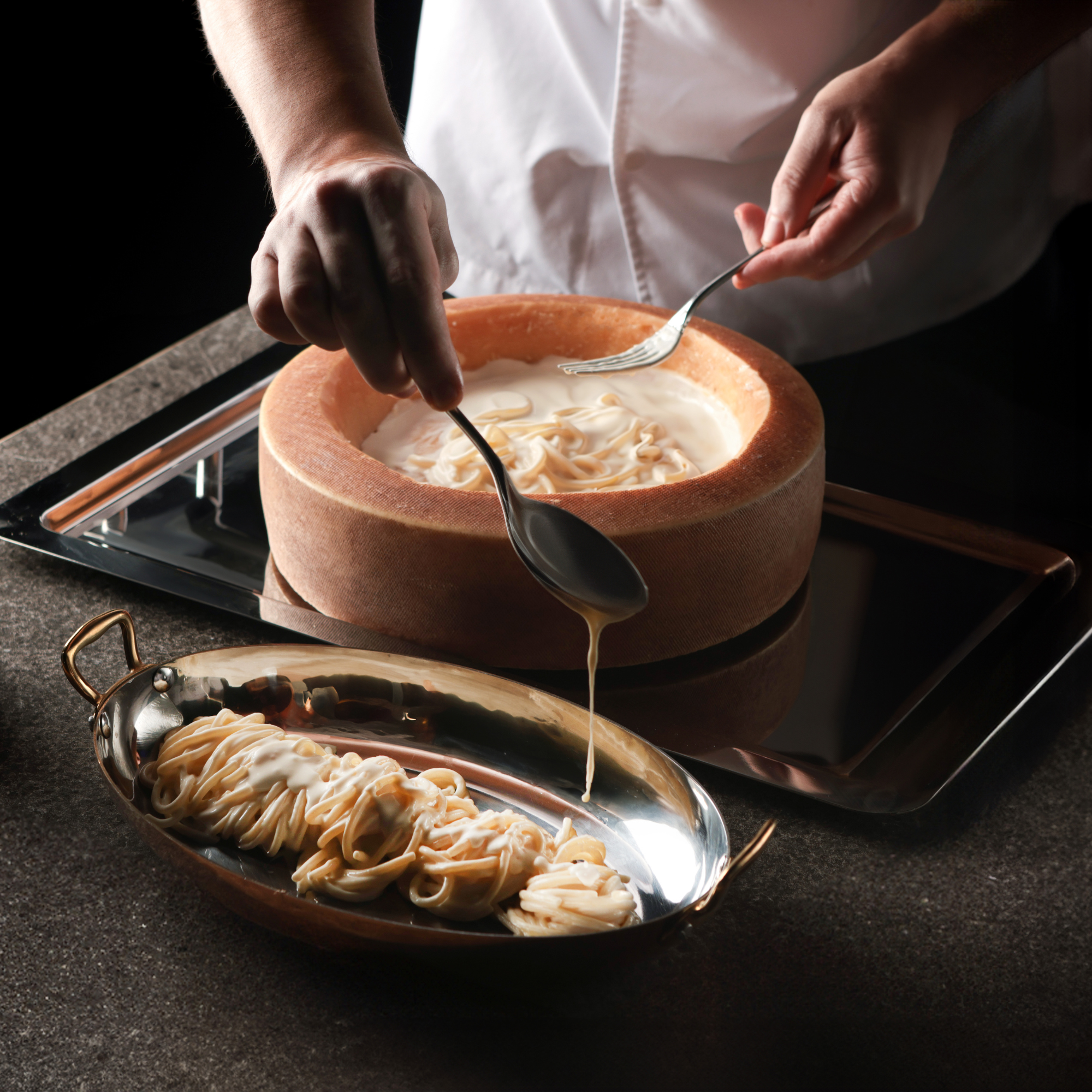 Fresh pasta finished inside a parmesan wheel with creamy sauce being poured over plated pasta.